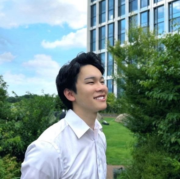 Smiling person in a white shirt standing outdoors near a modern building and greenery.