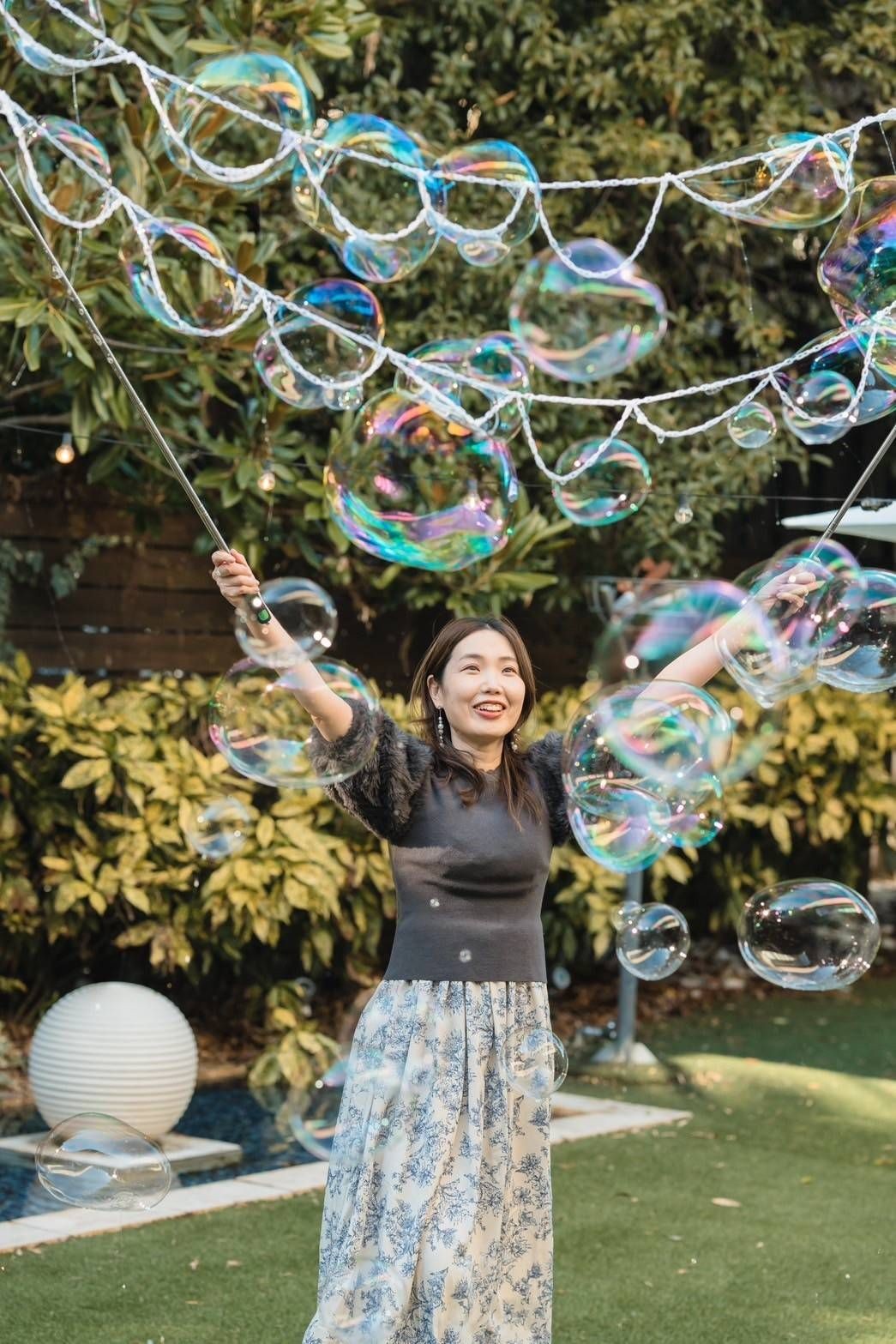 Woman joyfully creating giant soap bubbles outdoors, surrounded by greenery and plants.