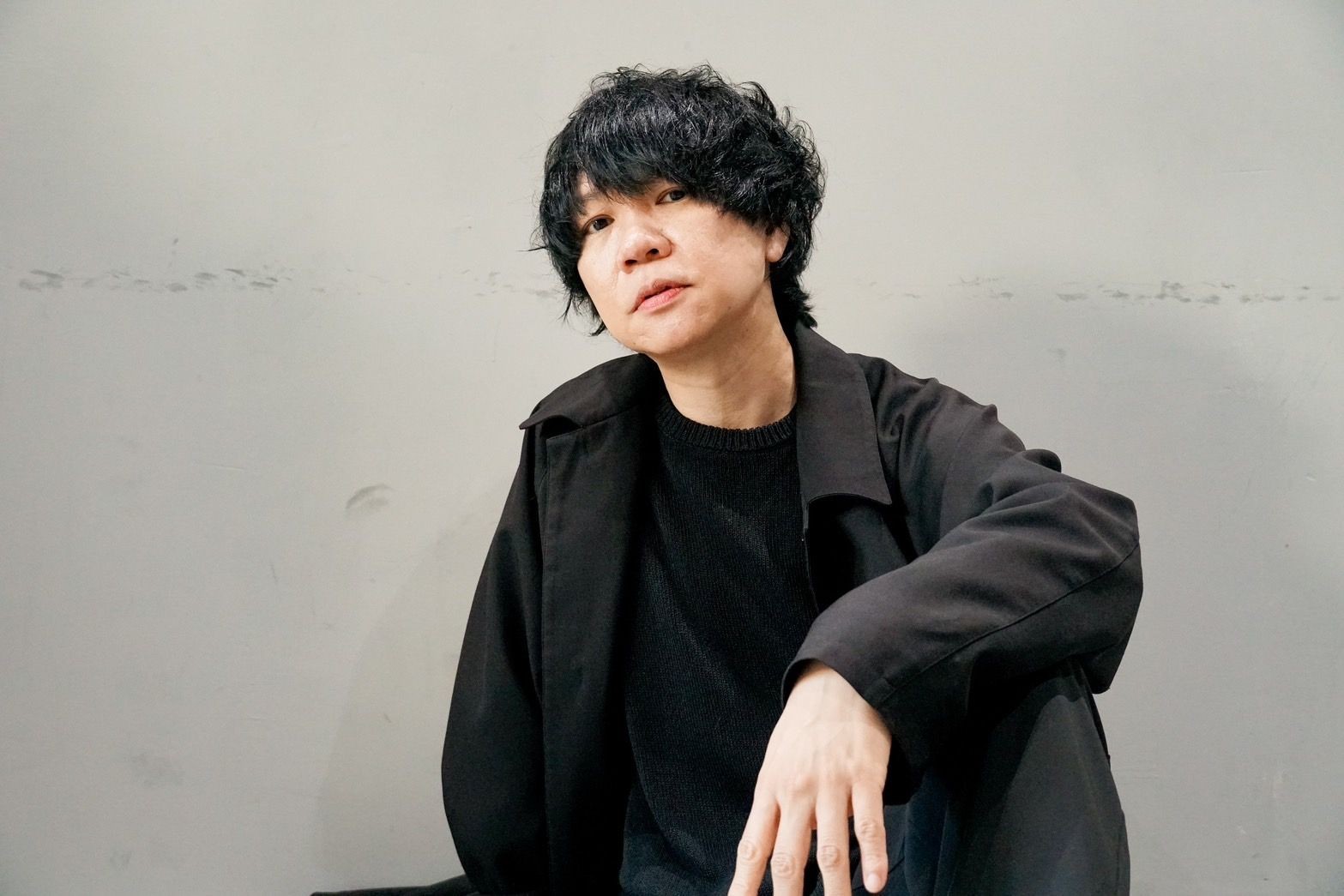 Person with black hair in a dark outfit sitting against a plain background, looking towards the camera.